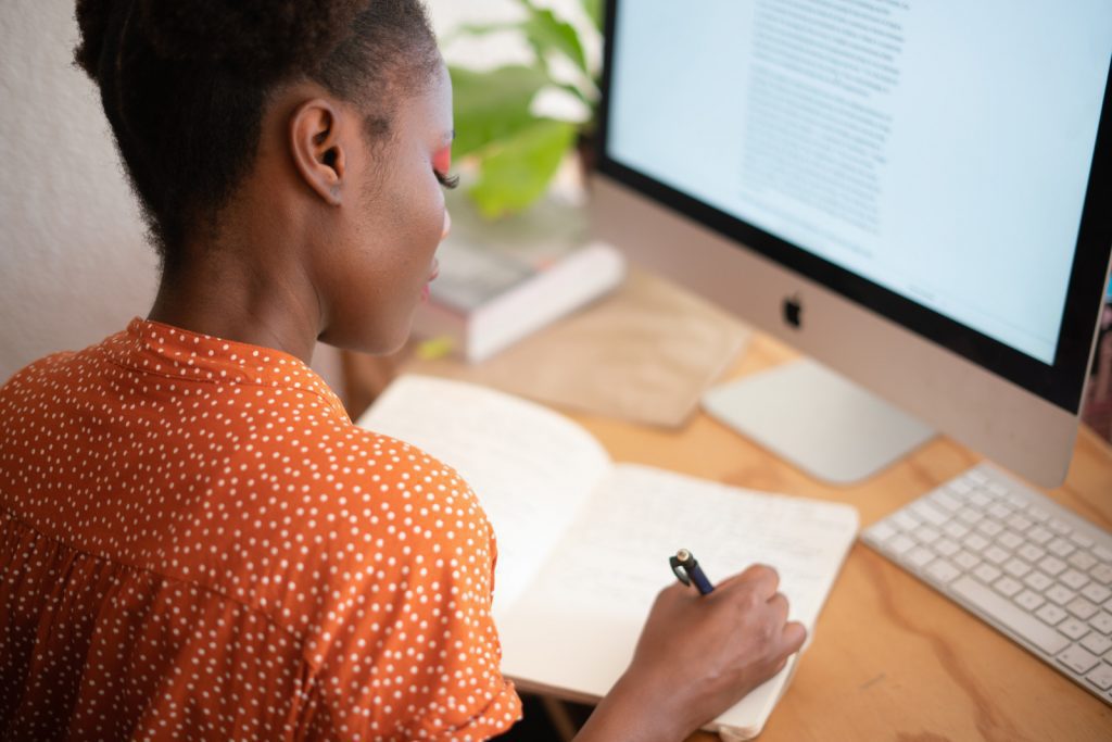 Woman writing in a notebook at a desk in front of a computer.