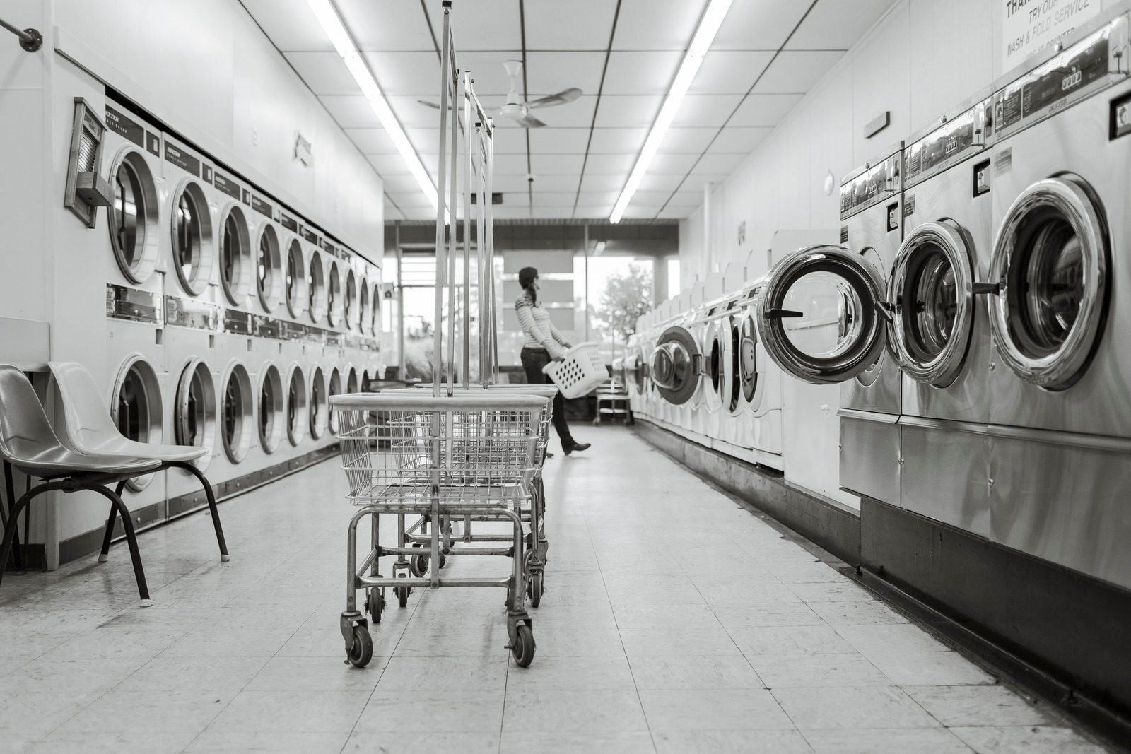 Grayscale photo showing metal laundromat carts at the center and machines on the side. A figure is in soft focus in the back carrying a basket.