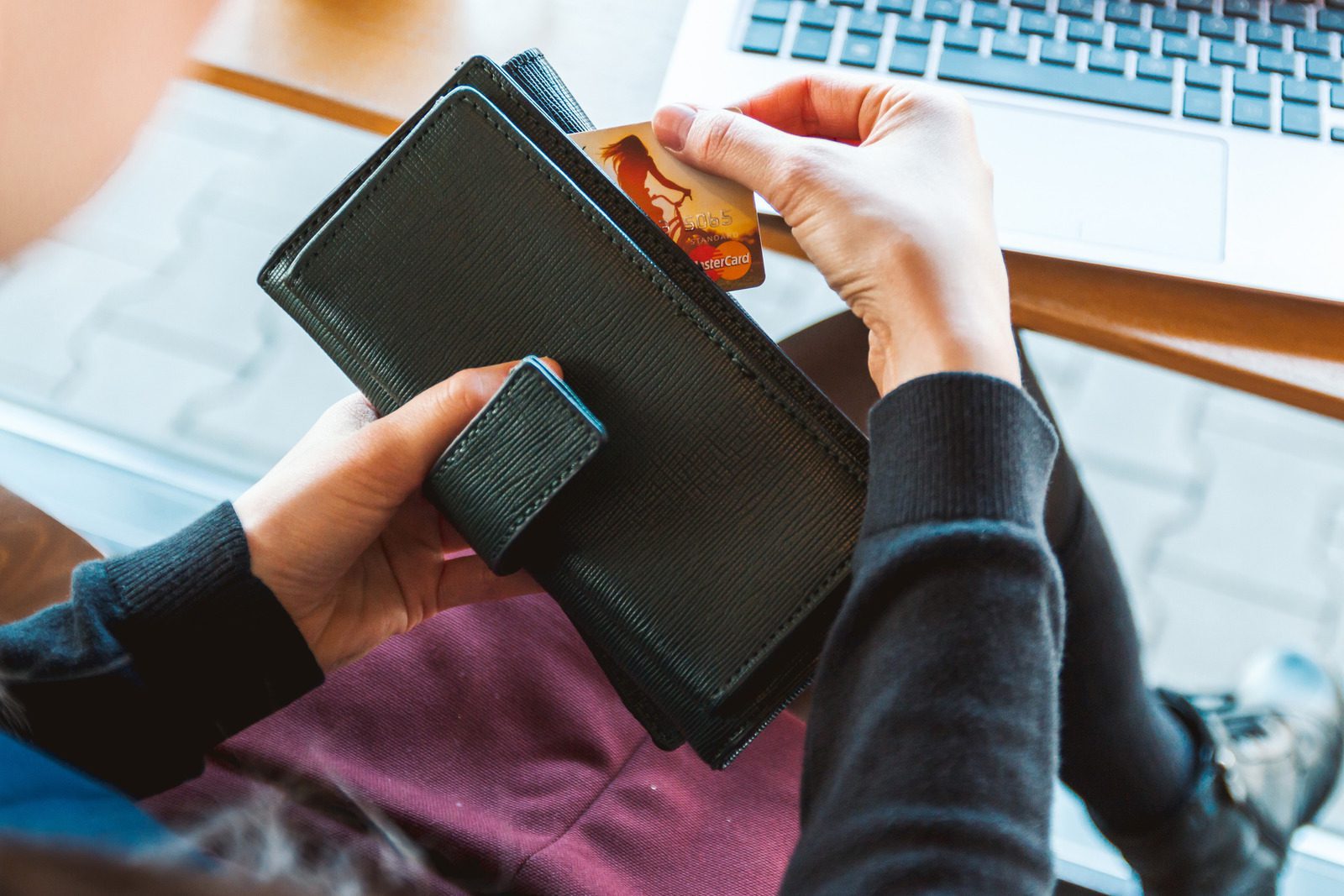 image of a woman's hands holding a black wallet with one hand pulling out a credit card and a laptop keyboard sits on a coffee table behind the hands