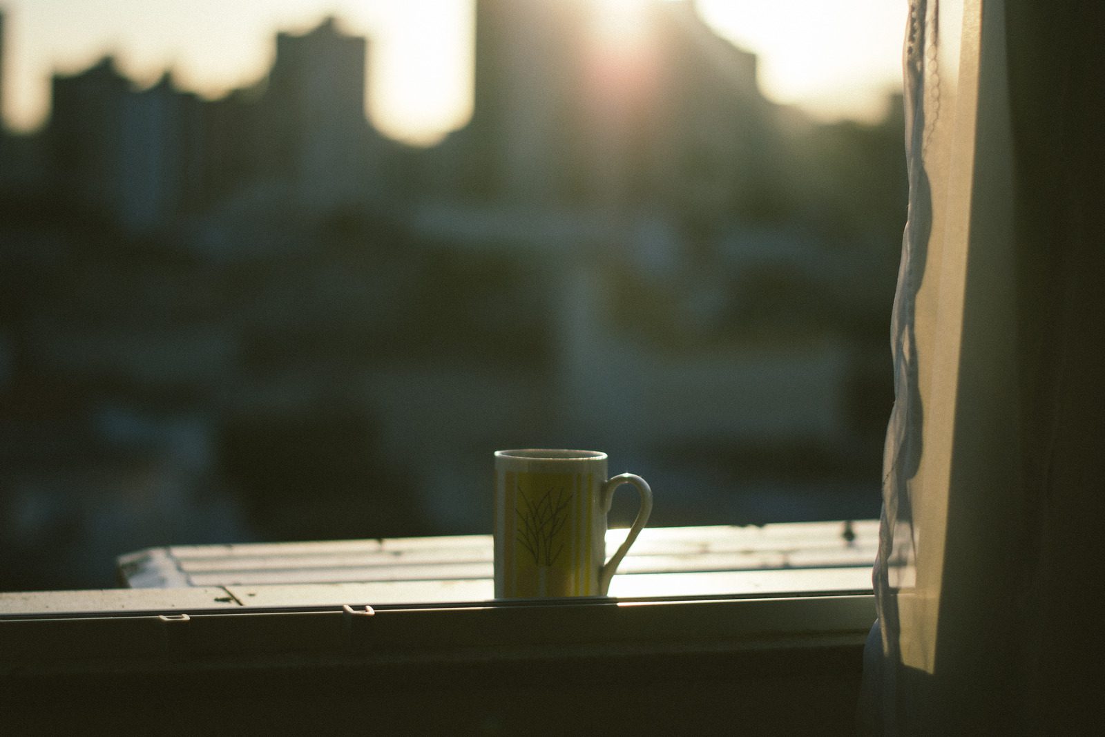 a mug sits in front of an open window with a curtain on the side and a city in blur in the background