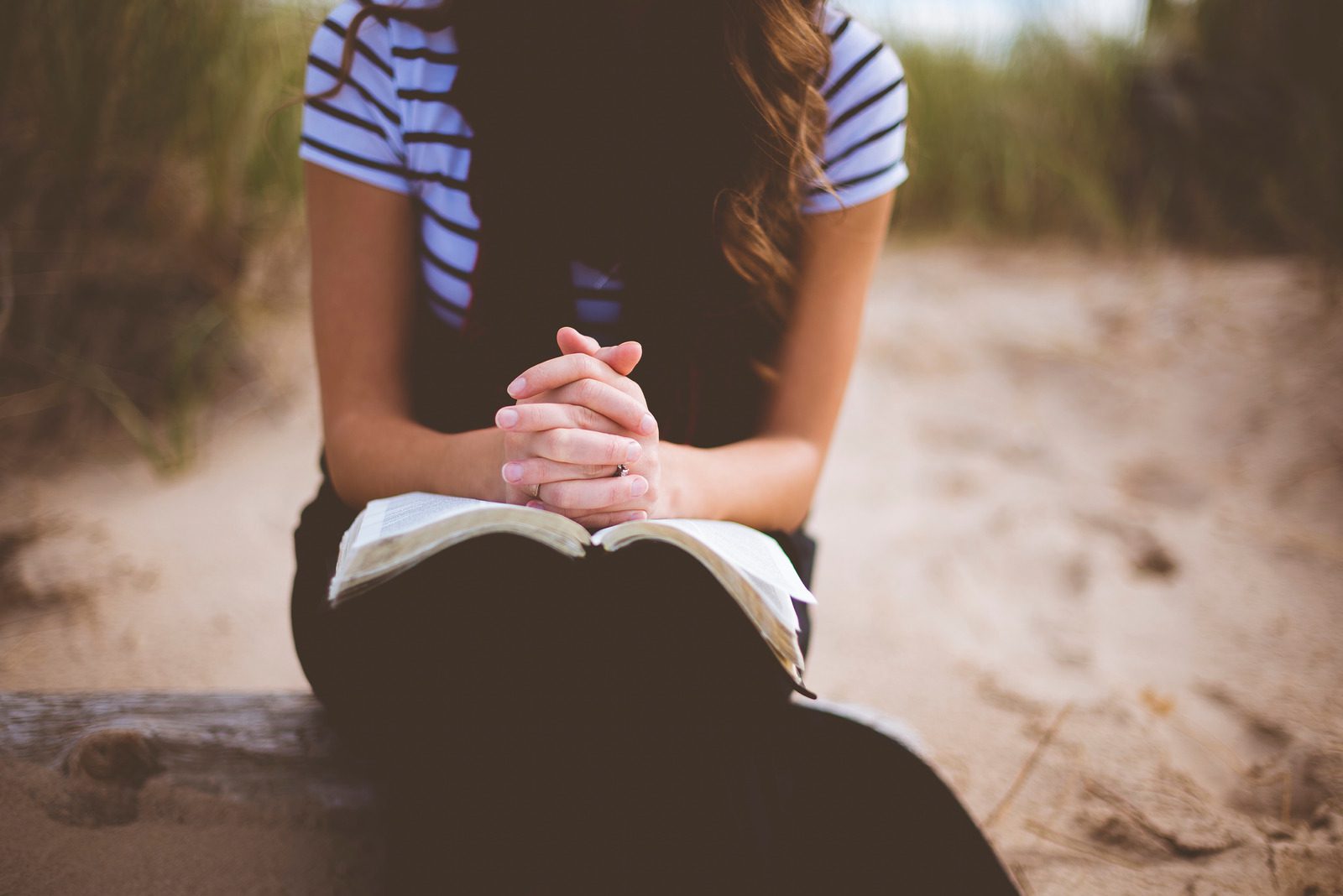 Soft focus image of a woman with praying hands resting on an open Bible. Behind her is a beach scene with sand and sea grass.