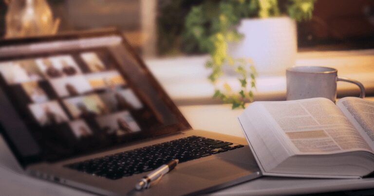 A laptop is open with a Zoom meeting showing multiple people. A Bible is open next to the laptop with a pen nearby.