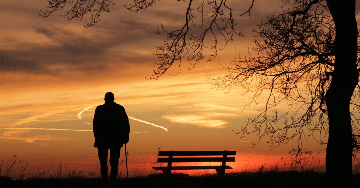 A man with a cane walks alone at sunset.