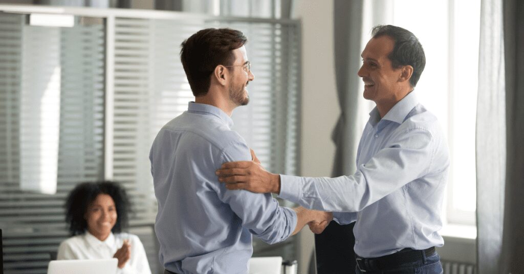 Two men shaking hands in an office with a woman seated near them.