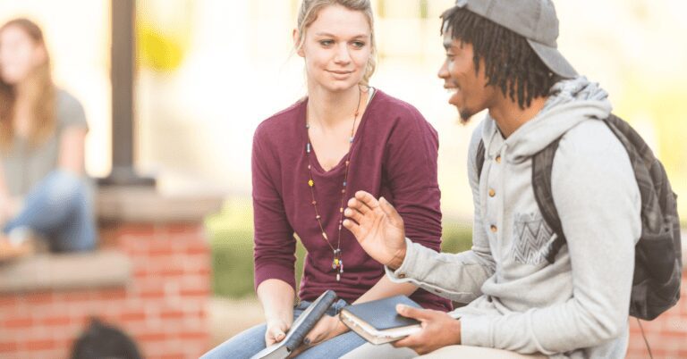 Two college students sitting on a brick wall talking while holding Bibles.