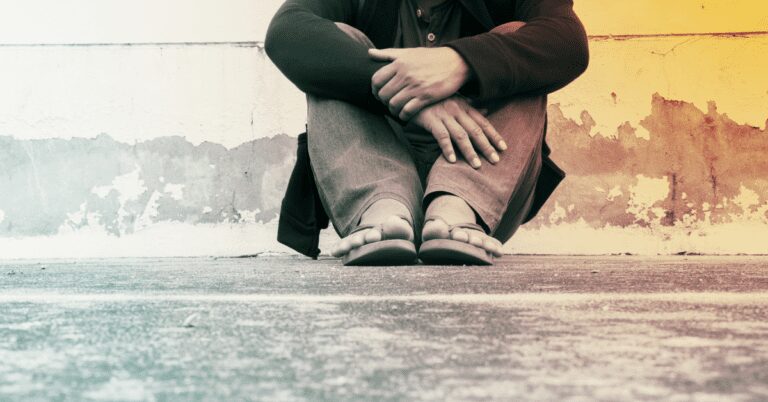 Person seen from waist down sitting against a wall clutching legs and wearing sandals.