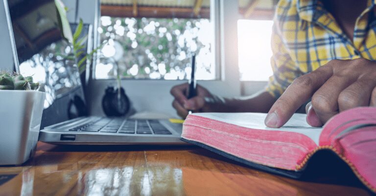 Closeup of man reading a Bible and taking notes next to a computer