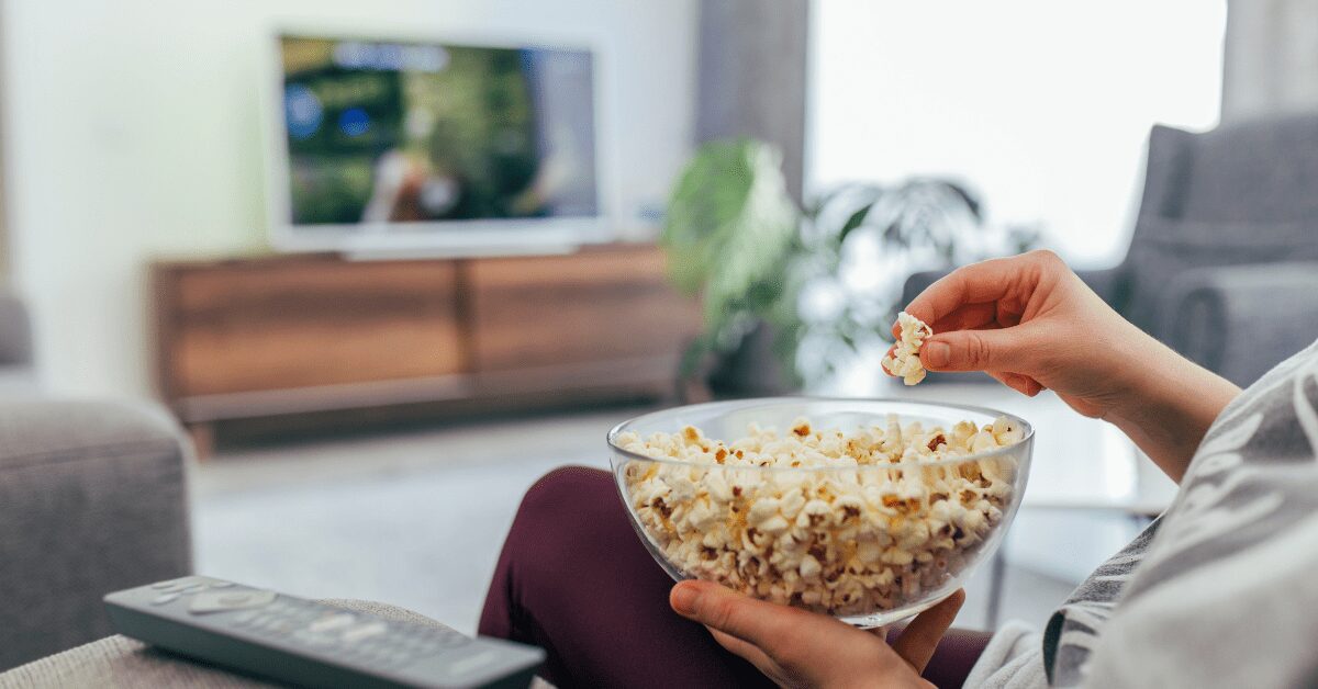 Closeup of hand holding popcorn bowl as a person sits on a couch watching TV.