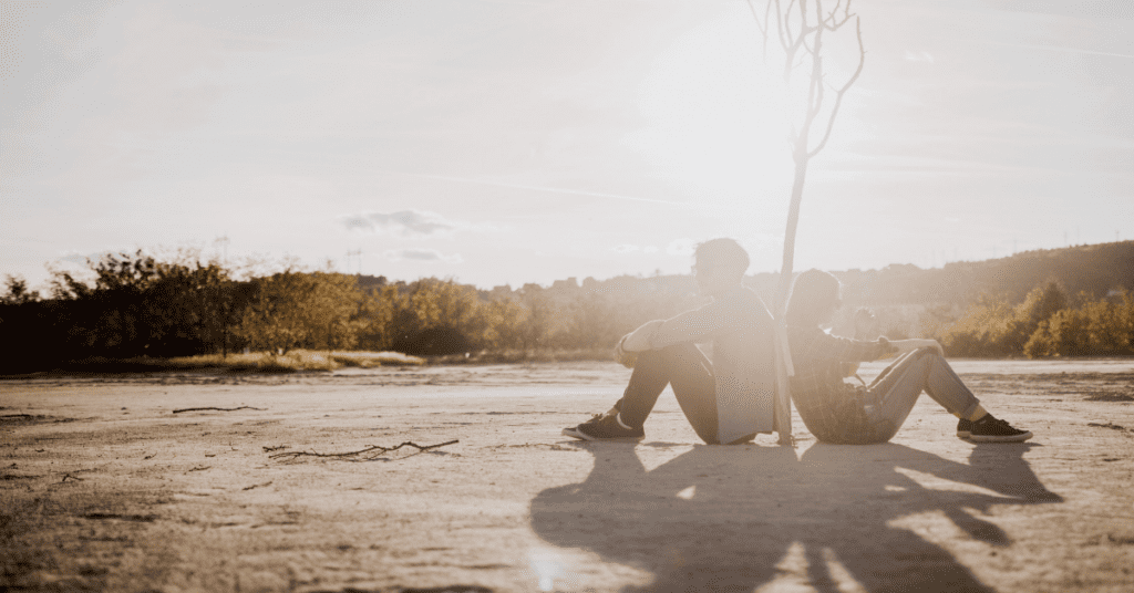 man and woman sitting back to back in the sun by a small tree