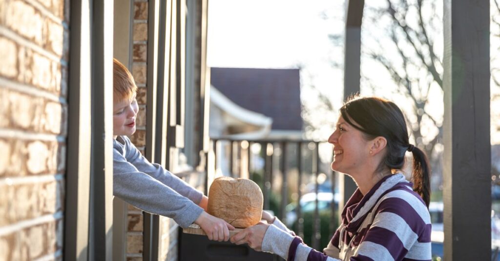 woman bent on her knees, passing a brown bag and book to a young boy in a doorway