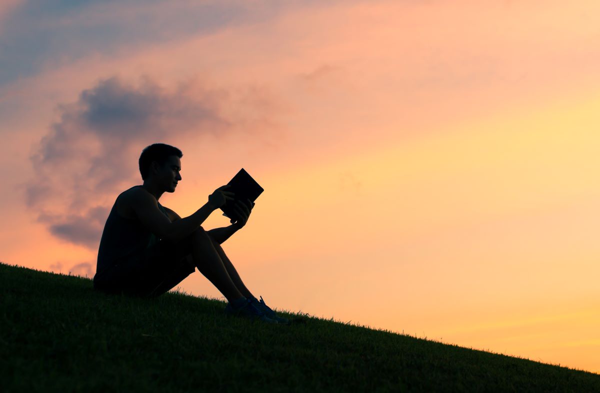 Young man sits on a hill reading the Bible with a sunset in the background.