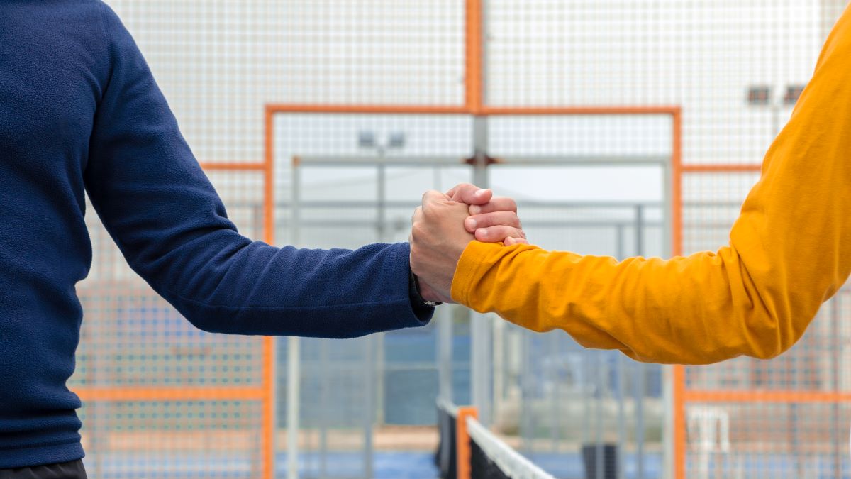 Two people, one in a blue shirt and the other in a yellow shirt, grasp hands across a net on a tennis court.