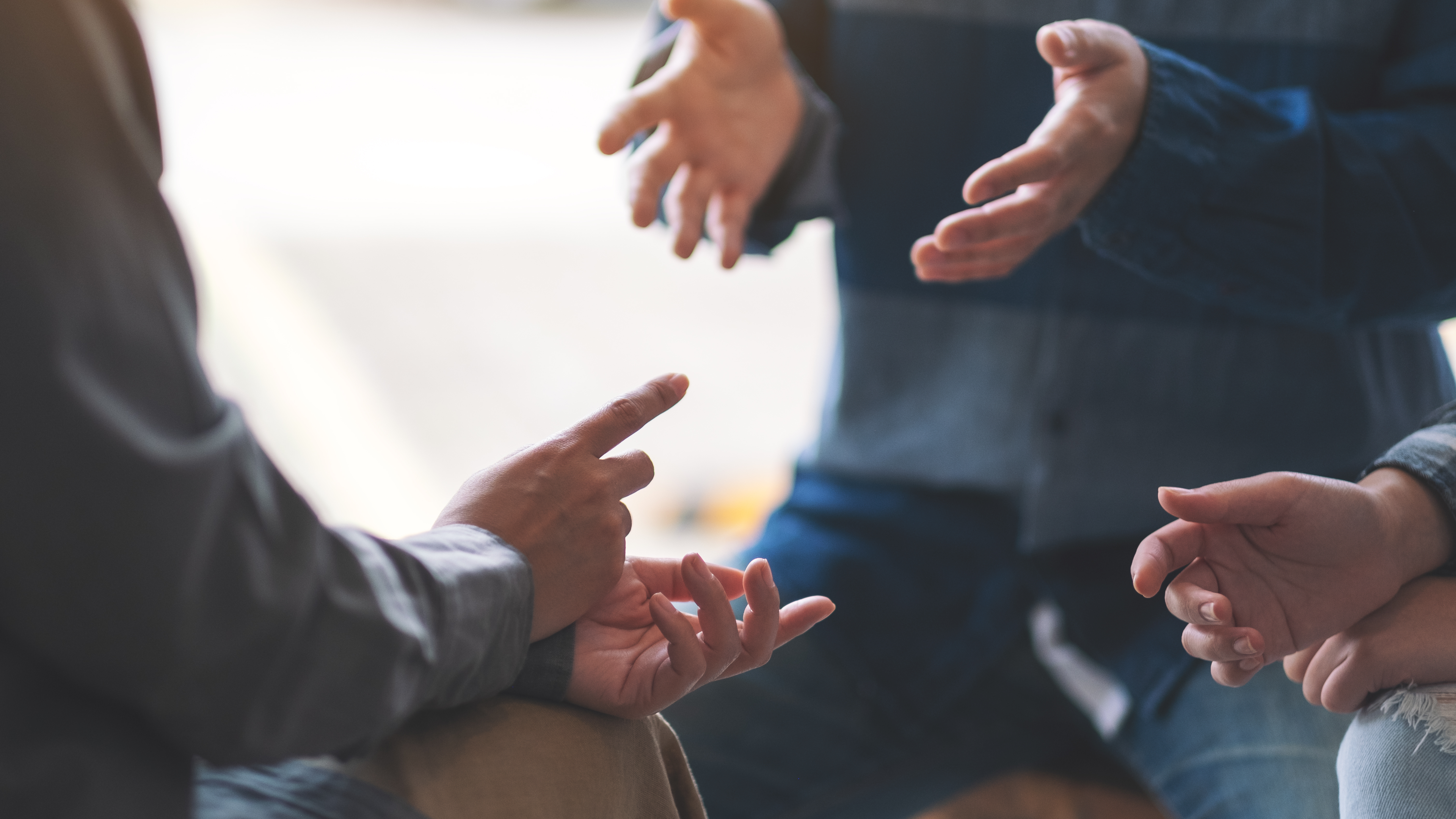 Image of three people sitting around in a group with their hands out, resting on their legs as if in conversation.