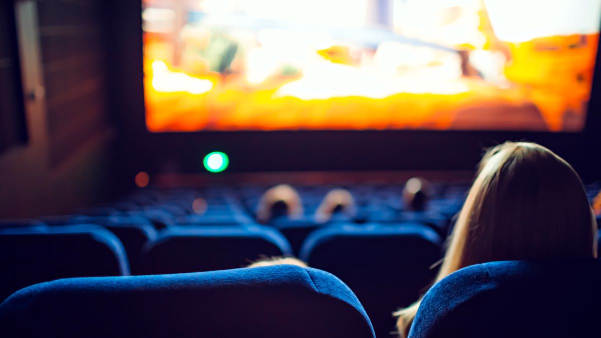 View from behind a moviegoer, including backlit, stadium-style seating and a bright, large movie screen.