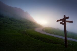 Faith+Lead | The sun crests over a winding road on a misty hillside; in the foreground is a directional signpost with planks of wood indicating distances and directions from the base of the hill.