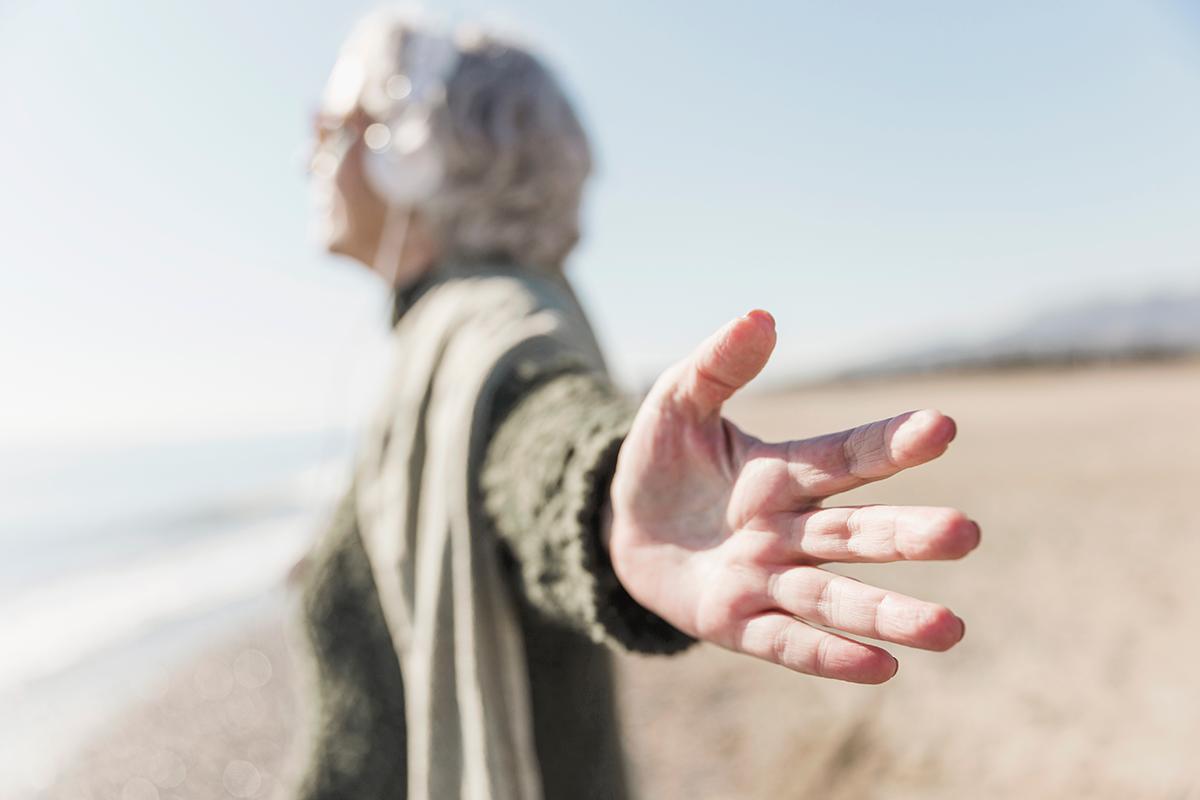 An older woman stretches out her hand toward the camera while standing on a beach, with the ocean and sky in the background.