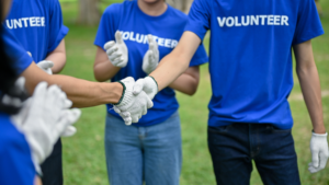 Volunteers in blue shirts shake hands while wearing work gloves, with others clapping in the background, symbolizing teamwork and collaboration.