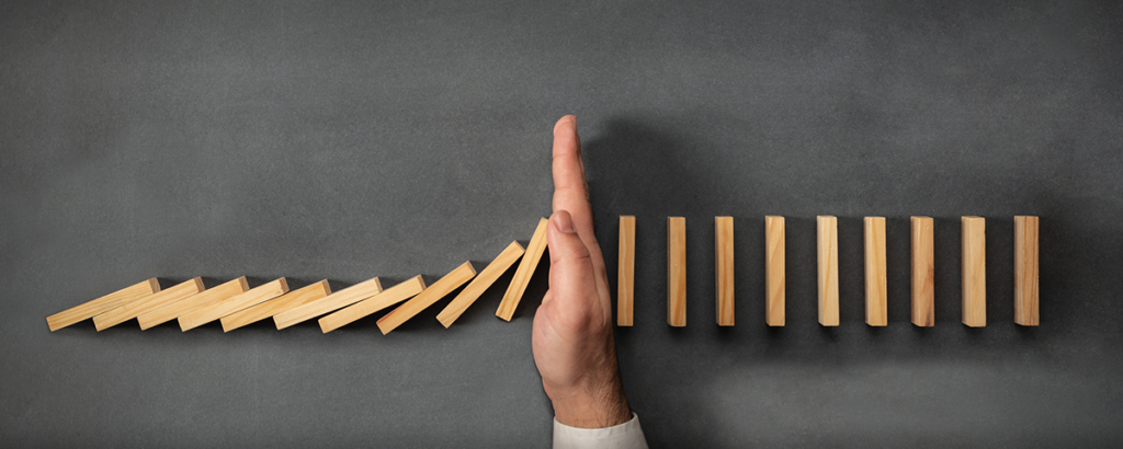 A hand stops a line of falling wooden dominoes on a dark surface, preventing them from toppling the remaining pieces.