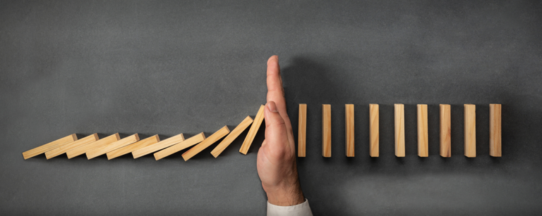 A hand stops a line of falling wooden dominoes on a dark surface, preventing them from toppling the remaining pieces.