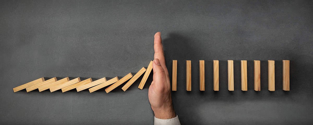 A hand stops a line of falling wooden dominoes on a dark surface, preventing them from toppling the remaining pieces.