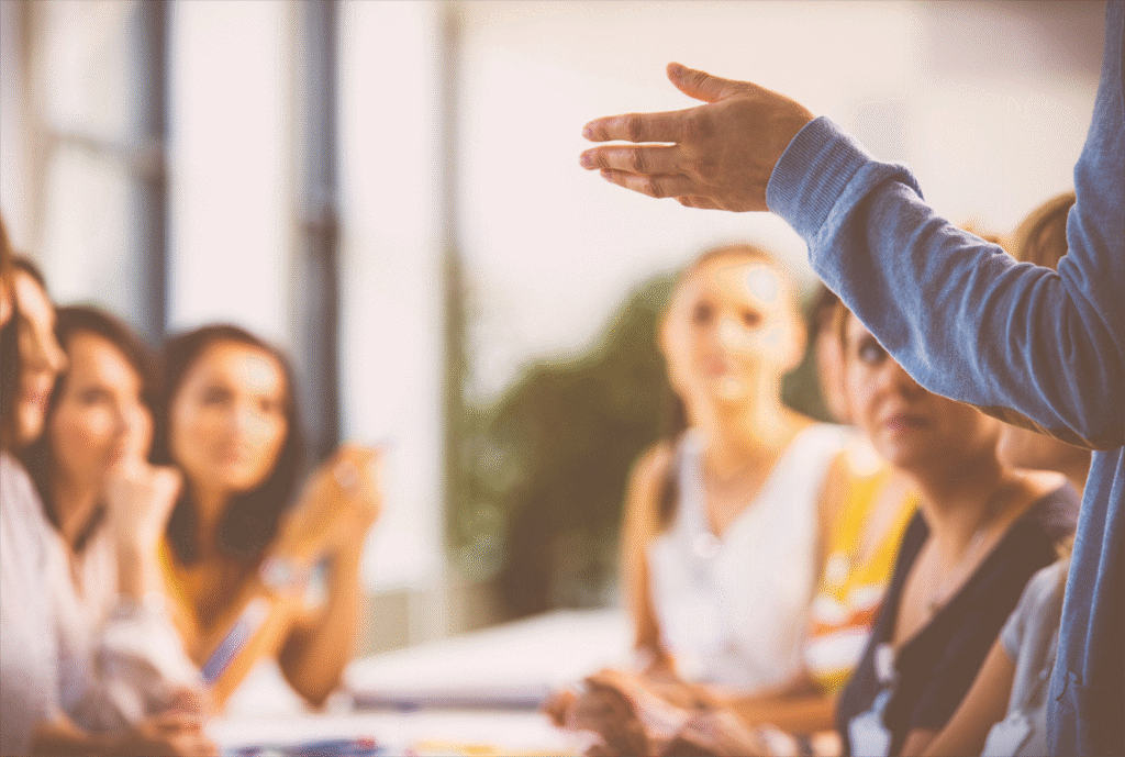 A person gestures while speaking to a group of attentive women seated at a table in a bright room.