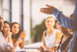 A person gestures while speaking to a group of attentive women seated at a table in a bright room.
