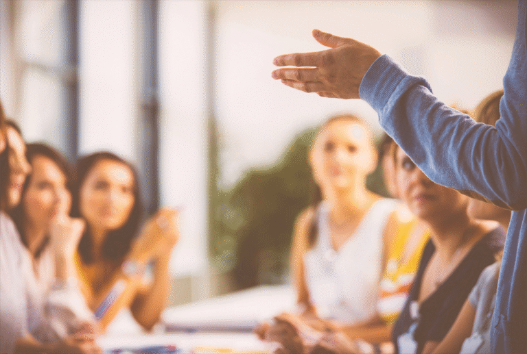 A person gestures while speaking to a group of attentive women seated at a table in a bright room.