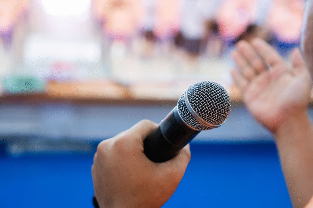 A person holds a microphone while speaking, with a blurred audience in the background.
