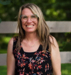 A woman with glasses and a floral shirt poses in front of a fence.