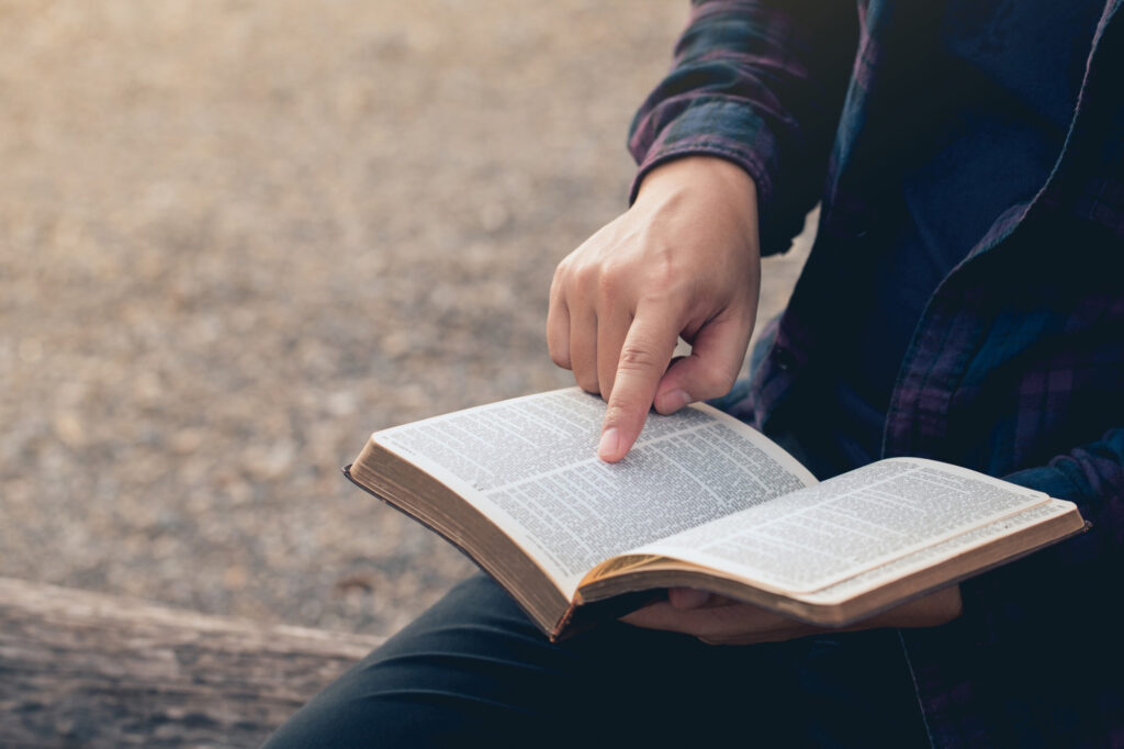 Person reading an open book outdoors, pointing at the text.