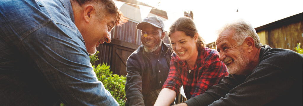 A group of four smiling adults, two men leaning in from the left and right and a Black man and a white woman in the middle, looking down and working together outdoors in a sunny, possibly rural or community garden setting.