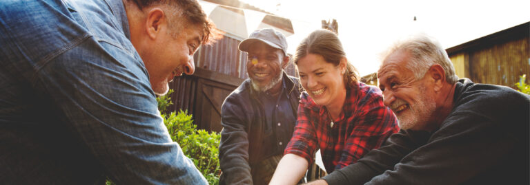 A group of four smiling adults, two men leaning in from the left and right and a Black man and a white woman in the middle, looking down and working together outdoors in a sunny, possibly rural or community garden setting.