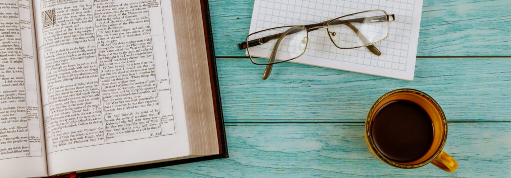 Open book on a turquoise wooden surface, with a notebook, reading glasses, and a cup of coffee.