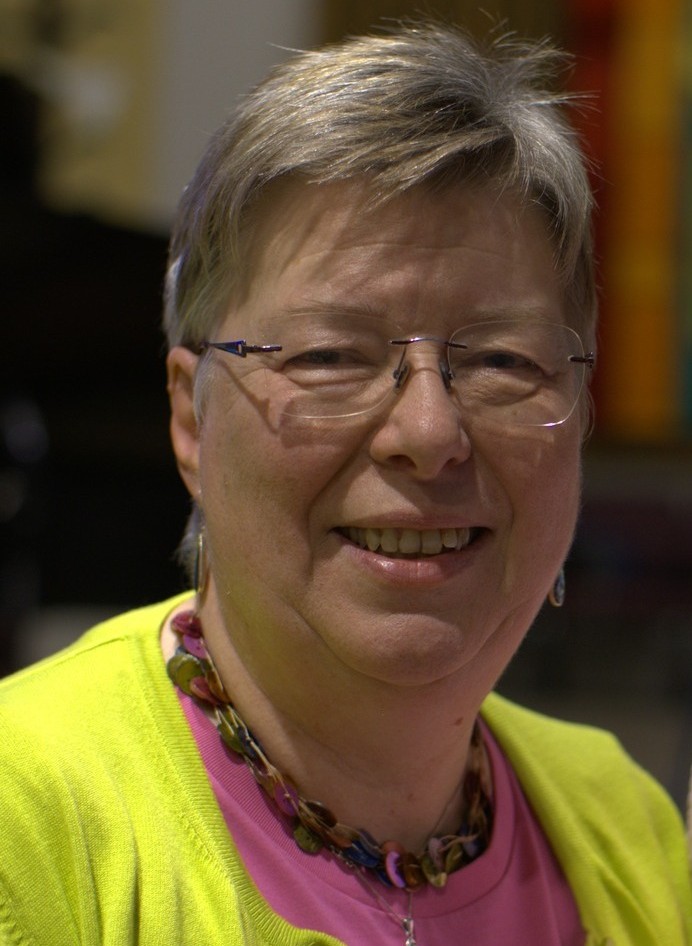 A smiling middle-aged woman with short, light-colored hair and glasses, wearing a bright lime green cardigan and a pink top with a beaded necklace.