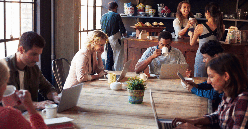 A busy, sunlit café where diverse people are working on laptops and drinking coffee at a large communal wooden table.