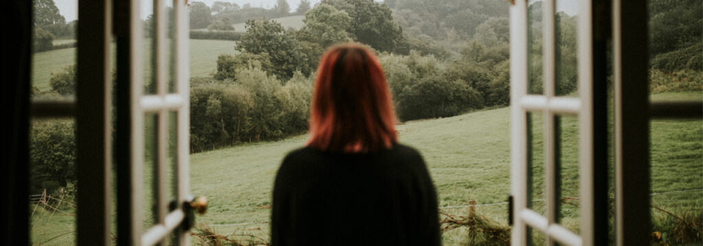 A wide, soft-focus view from behind a woman with shoulder-length red hair as she looks out of an open white-framed window onto misty, rolling green hills and dense woodlands.