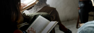 An overhead, close-up shot of a small group of people sitting in a circle, focused on reading open books together. The lighting is soft and moody, emphasizing the pages of the books and the hands of the participants. The atmosphere suggests a quiet, communal moment of study or reflection.