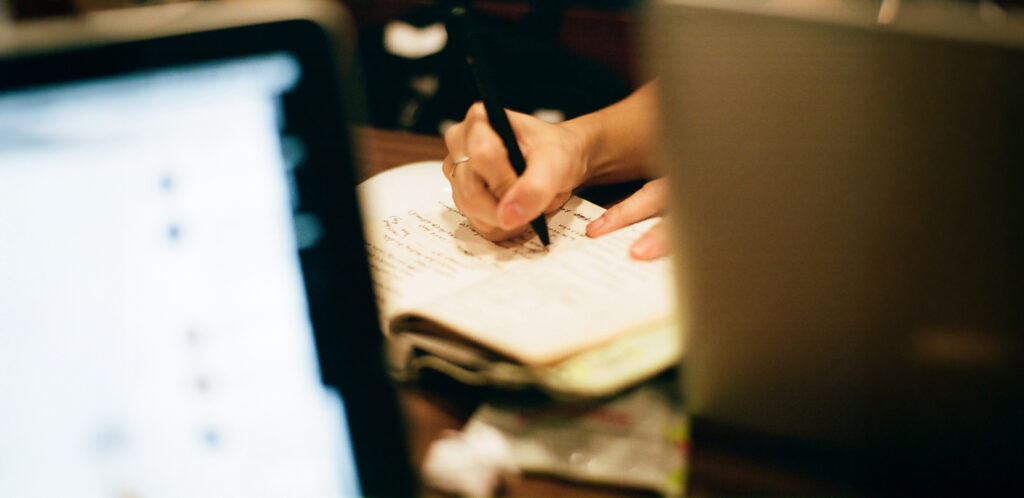 A close-up of a person's hand writing in a notebook with a black pen, framed between two blurred laptop screens in a dimly lit setting.