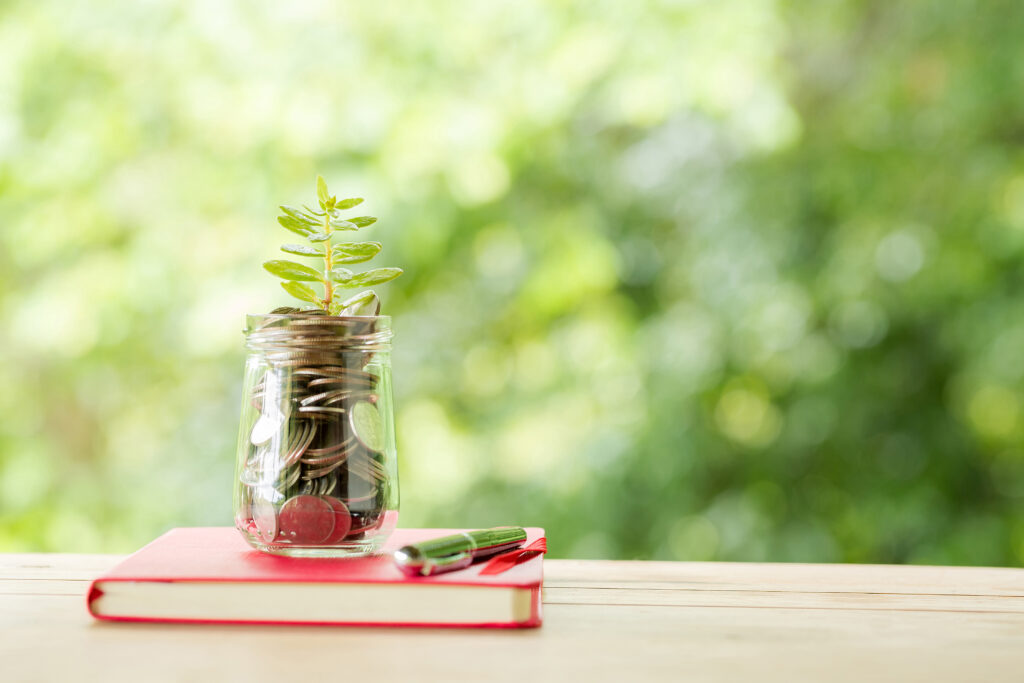 A small green plant growing out of a glass jar filled with coins, resting on a red notebook.