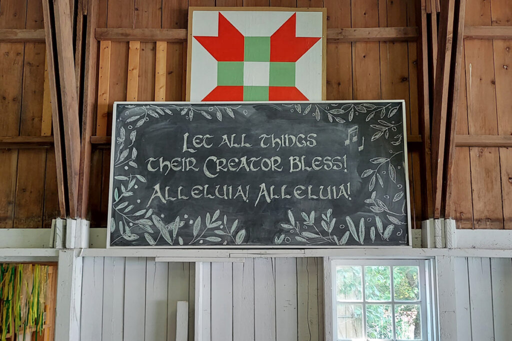 A chalkboard in a rustic barn with the hand-written text: "Let all things their creator bless! Alleluia! Alleluia!"