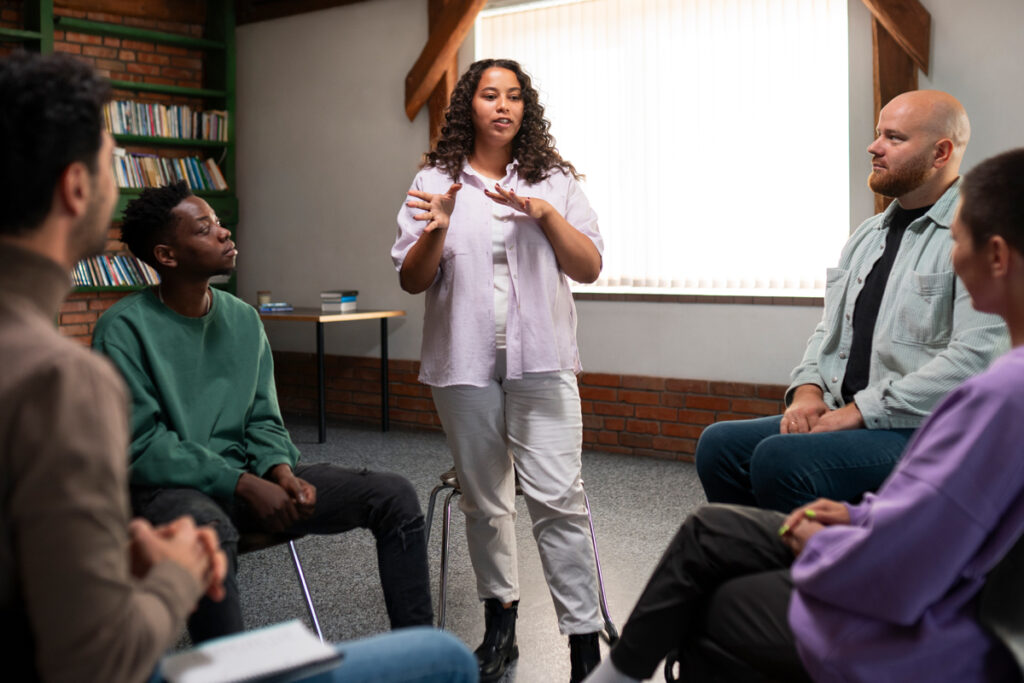 Gemini said A woman stands in the center of a seated circle of people, gesturing with her hands as she speaks to the group in a library or community room.