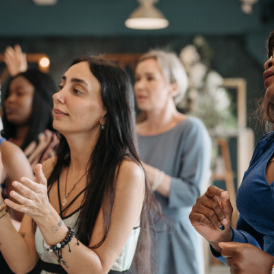 A candid, medium shot of a diverse group of women standing together and clapping in a brightly lit indoor space.