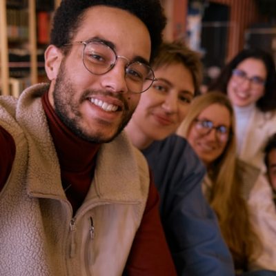 A smiling man with glasses takes a selfie with a group of friends indoors.