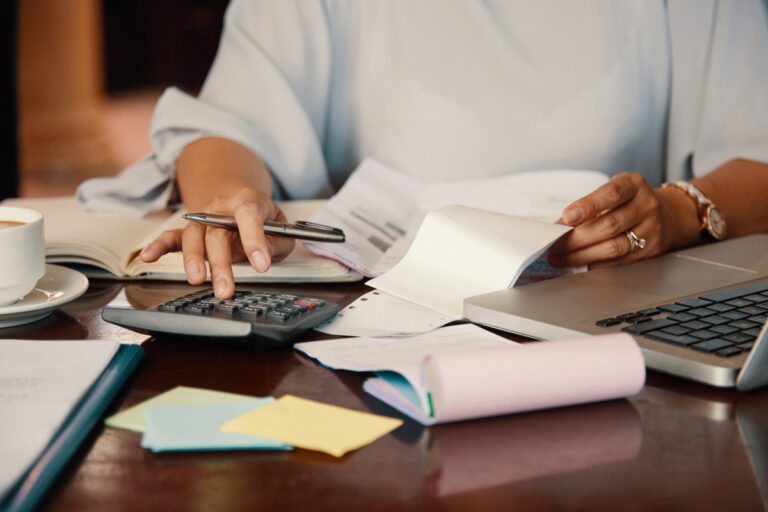 A person uses a calculator and reviews financial documents at a desk with a laptop and coffee.