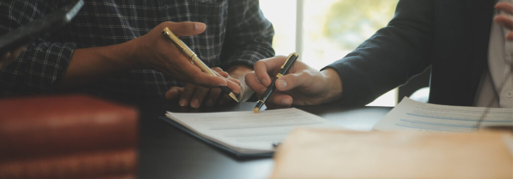 Two people at a desk review a document together, each holding a pen and pointing at the paperwork during a professional discussion.