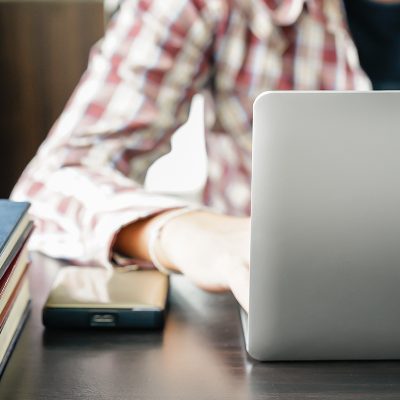 : A person in a plaid shirt working on a laptop at a desk with books and a portable hard drive.