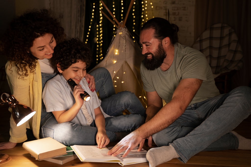 A family—a mother, father, and young son—smiling and reading a book together on the floor at night, illuminated by a flashlight and a small lamp near an indoor play tent with string lights.