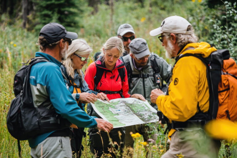 A group of six hikers gathered in a grassy, wooded area, looking closely at a map held open by a woman in a red jacket.