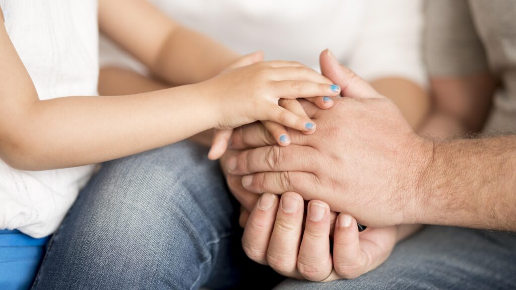 A close-up of a young child's small hand with blue nail polish resting gently on top of an older man's larger, weathered hands.