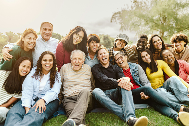 A large, diverse group of smiling, multi-generational people sitting and standing close together outdoors on a grassy field.